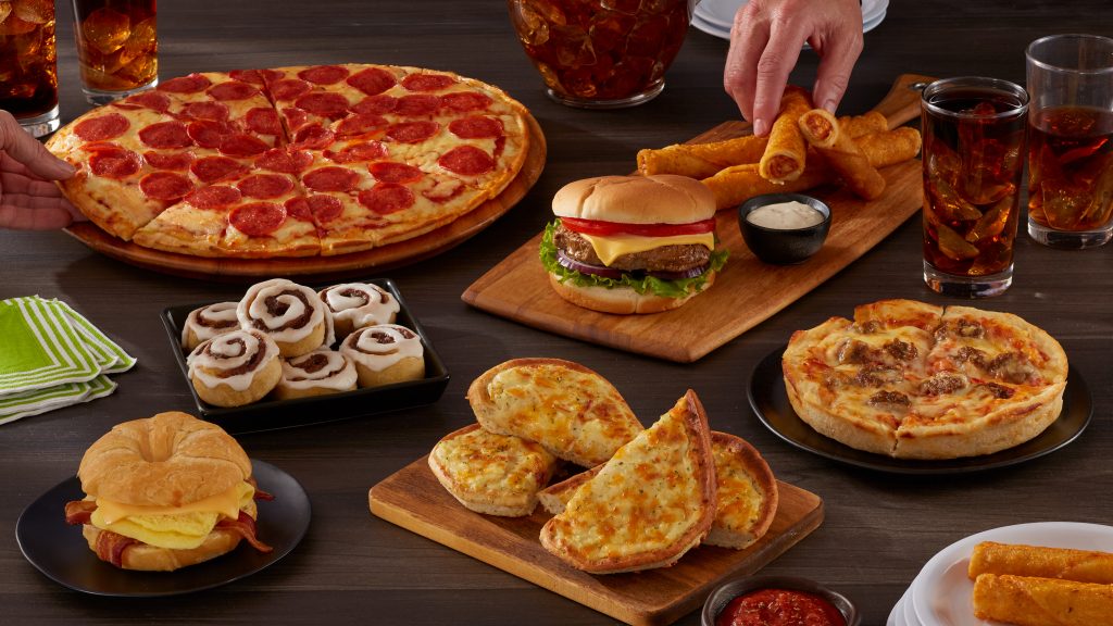 Assorted convenience food items including pepperoni pizza, breakfast sandwich, burger, breadsticks, taquitos, and cinnamon rolls arranged on a table.
