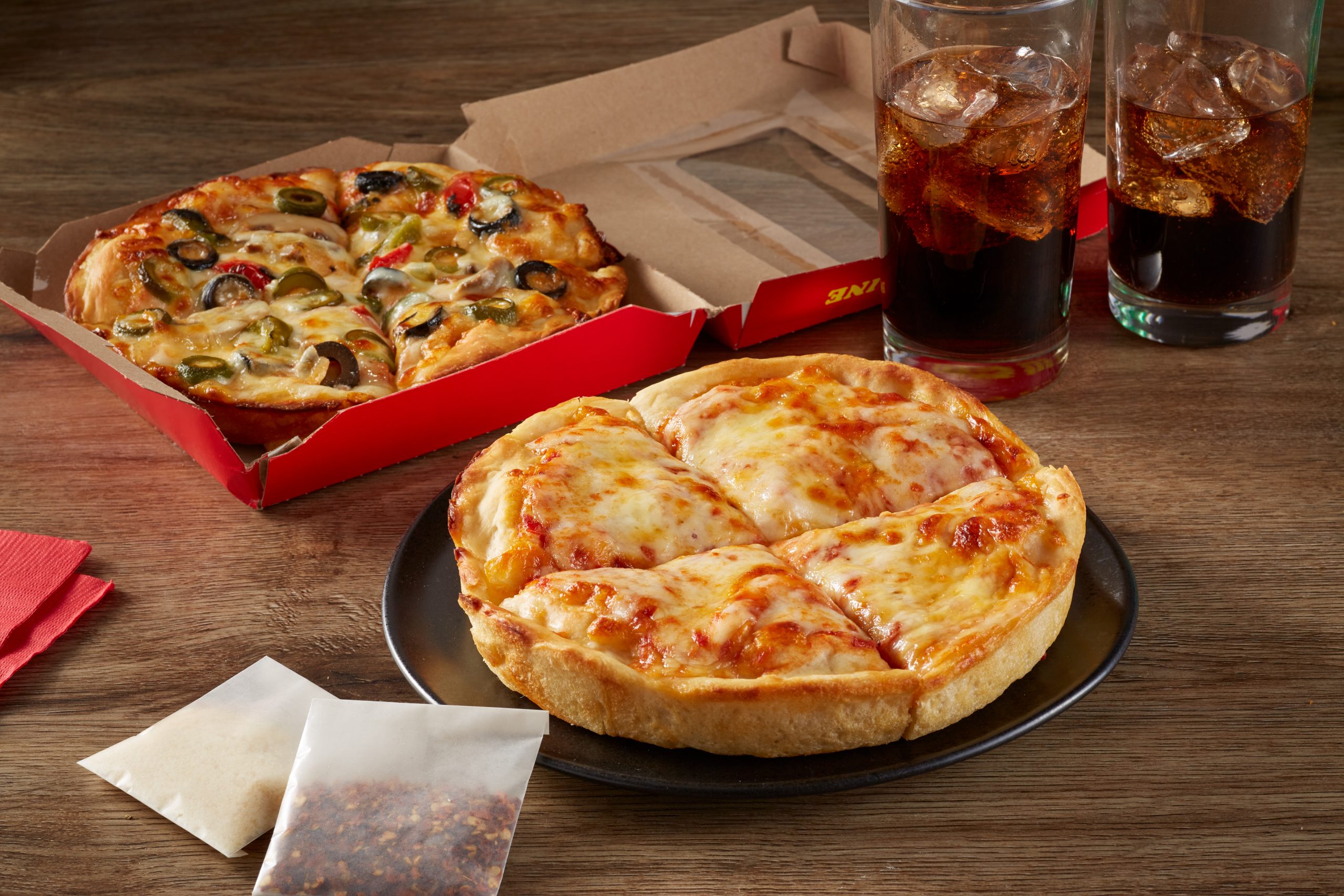Personal pan cheese pizza and veggie pizza served with fountain drinks and seasoning packets on a wooden table.
